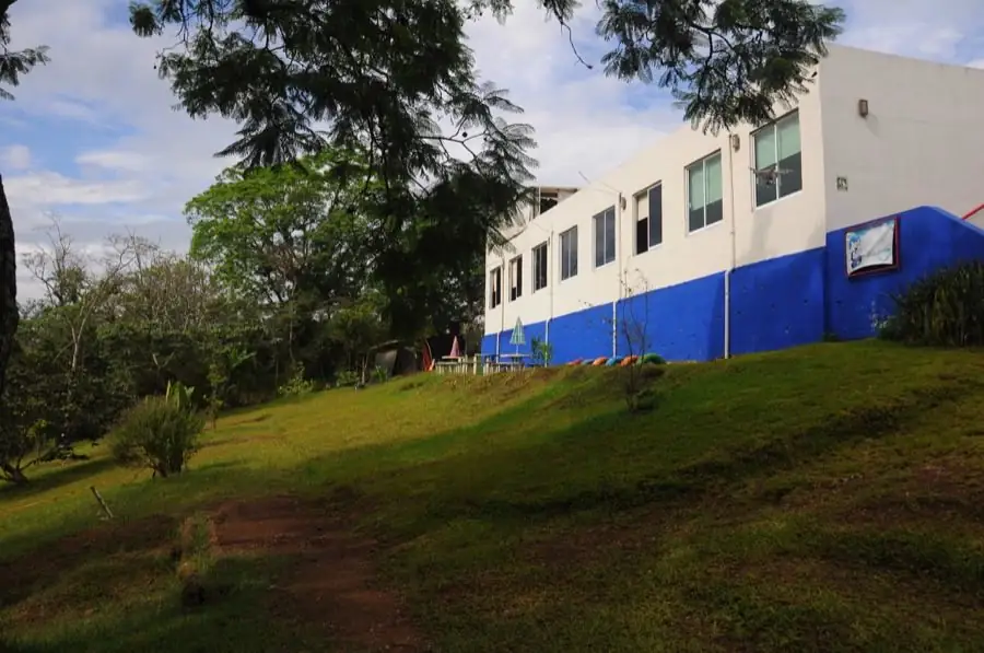 Edificio del colegio, blanco con franja azul, sobre una colina con pasto